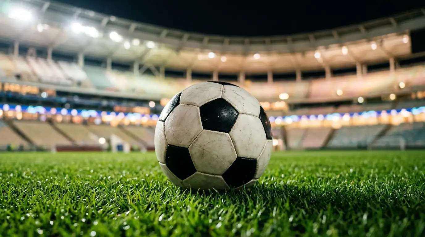 Ballon de football posé sur la pelouse d'un stade moderne sous les projecteurs, tribunes floues en arrière-plan
